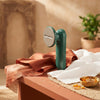 A green portable handheld garment steamer placed on a terracotta table next to white linen fabric in a modern home.