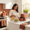 A South Asian woman using a 2-in-1 cooking oil spray bottle in a sunlit kitchen with terracotta and burnt sienna decor.